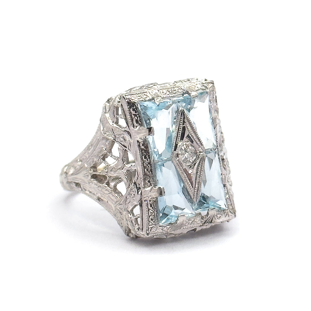 Off the hand now against plain white, the ring angled to show the engraved filigree shoulders, the openwork gallery beneath the four aquamarines, and prongs gripping the corners of the face.