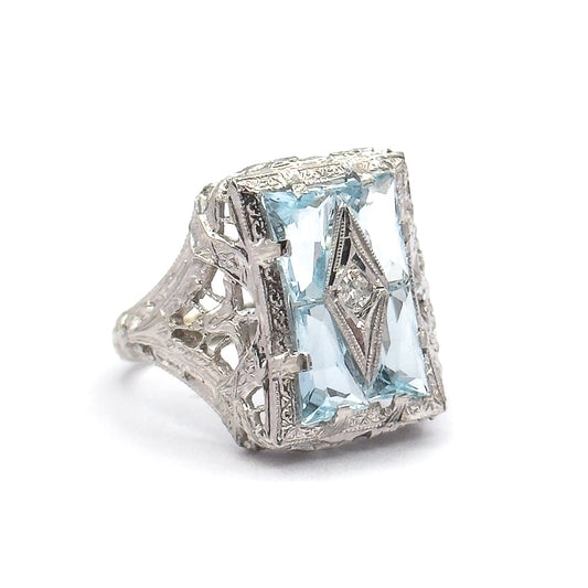 Off the hand now against plain white, the ring angled to show the engraved filigree shoulders, the openwork gallery beneath the four aquamarines, and prongs gripping the corners of the face.