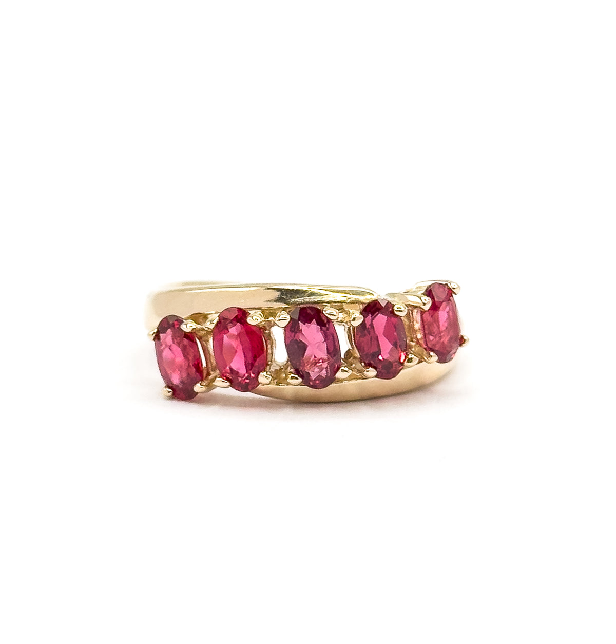 Close straight-on view against white with the ring low in the frame, the five oval red spinels and the prongs between them filling the face while the yellow gold shank curves away behind.