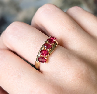 Close overhead view of the five prong-set oval red spinels running diagonally across the finger, the yellow gold face catching light as it narrows into the shank.