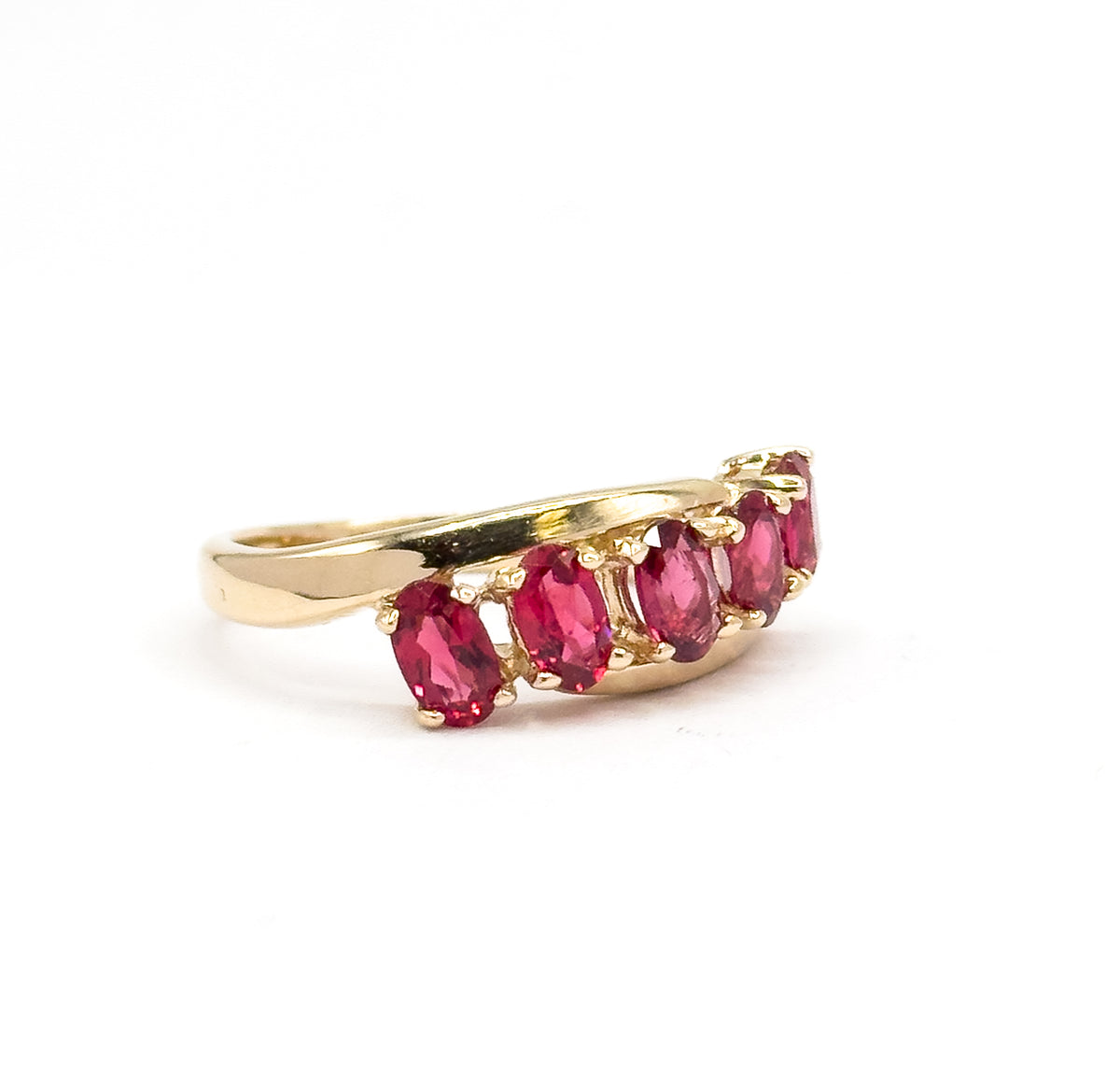 Low side-on view at eye level against white, where the yellow gold shank curves up into the stone row and the oval red spinels are seen edgewise, each gripped by side prongs.