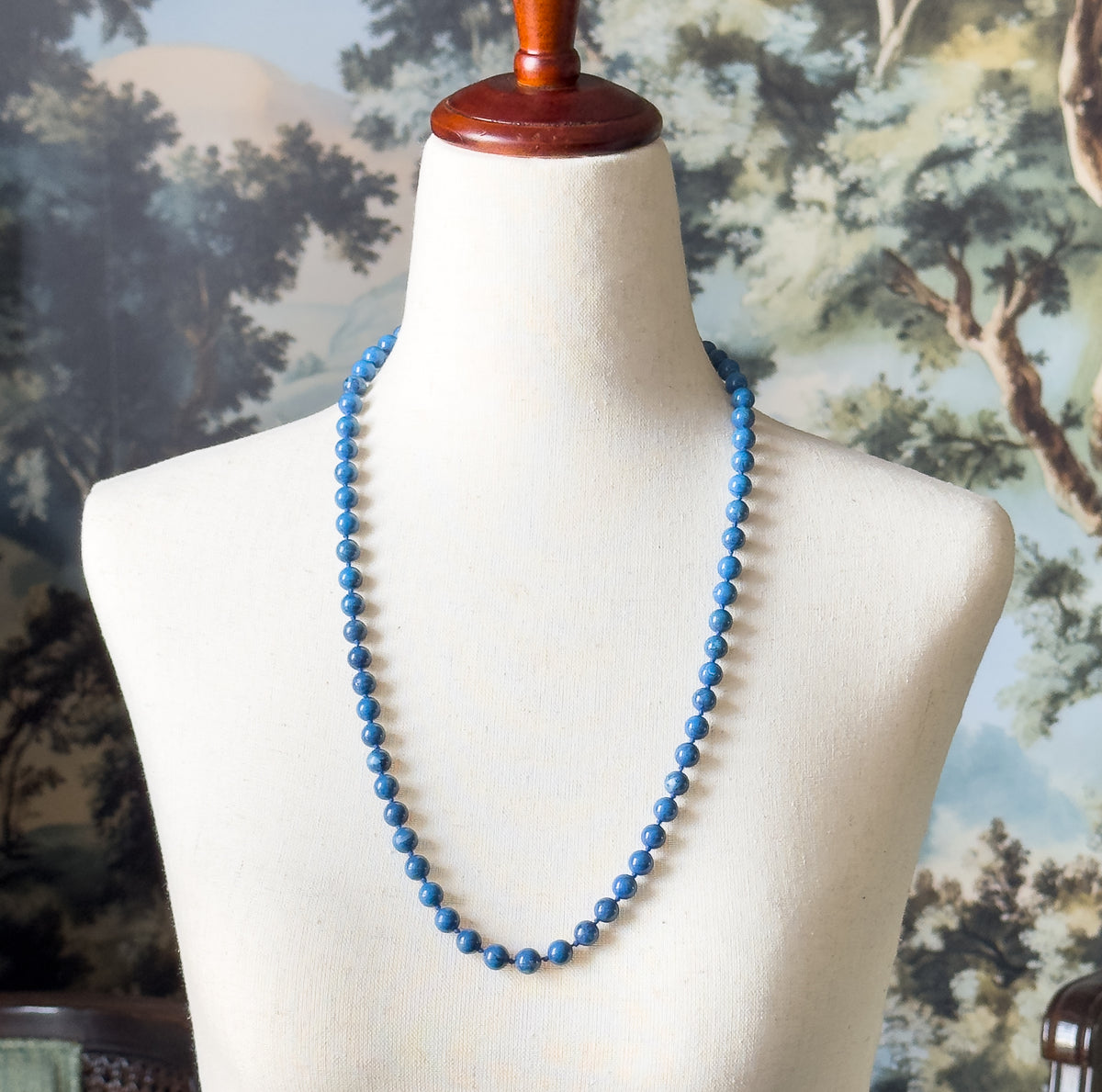 Shown at the neck on a cream dress form against a painted woodland backdrop, the single strand of round cobalt blue beads hangs in a long loop reaching the mid-chest.