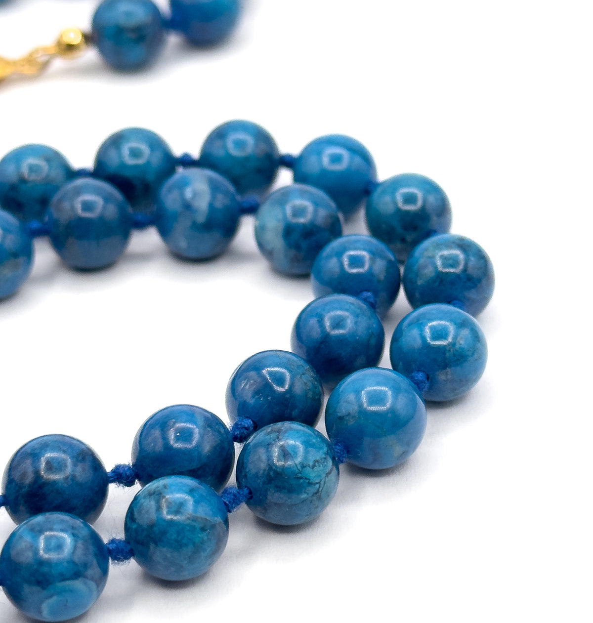 Close macro of the curving strand on white, showing the deep blue mottling within each polished bead, the blue knots between them, and the yellow-toned clasp blurred behind.