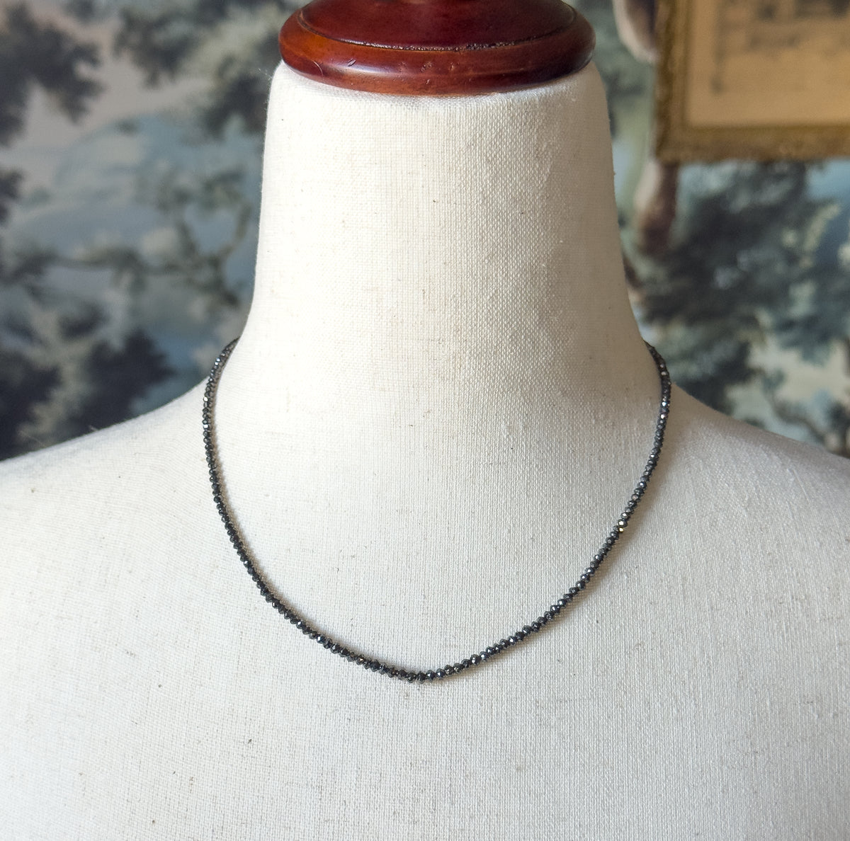 A black diamond bead necklace in 18K white gold, shown at the neck on a cream linen bust form, the graduated faceted beads tapering toward the centre of the strand.