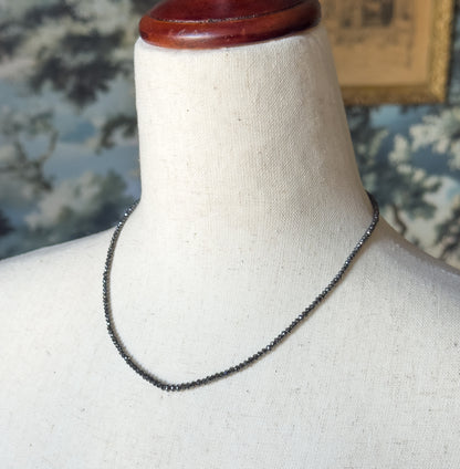 A closer view of the strand curving across the linen bust form, the small faceted black diamond beads catching light along the neckline against a blue floral backdrop.