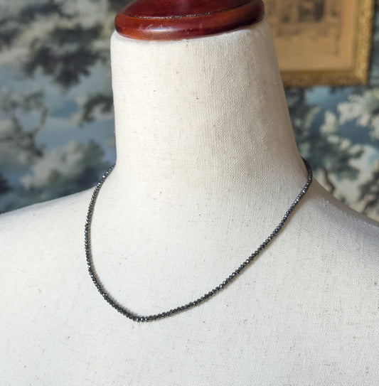 A closer view of the strand curving across the linen bust form, the small faceted black diamond beads catching light along the neckline against a blue floral backdrop.