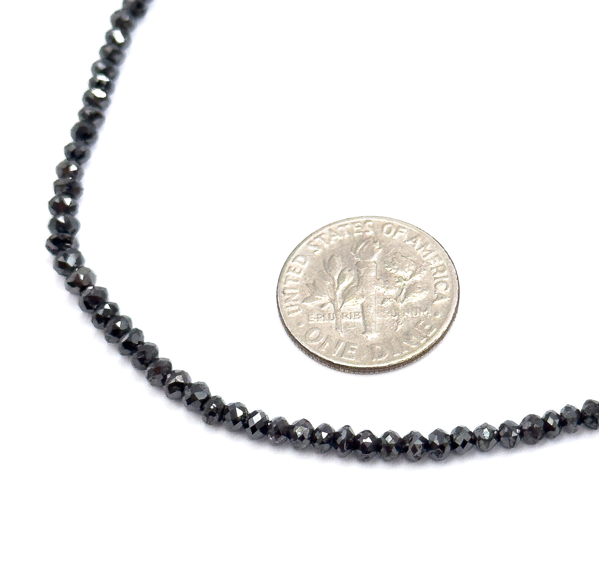 The strand laid in a curve on a white background beside a dime, the coin showing how small the graduated faceted black diamond beads are, each barely a fraction of its width.