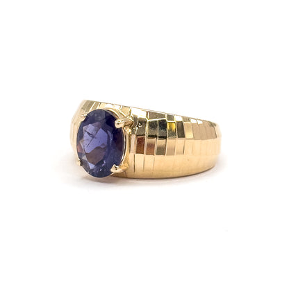 Three-quarter view with the oval iolite tilted toward the viewer so its deep bluish purple facets show between top and bottom prongs, the stepped faceted gold band rising away to the right.