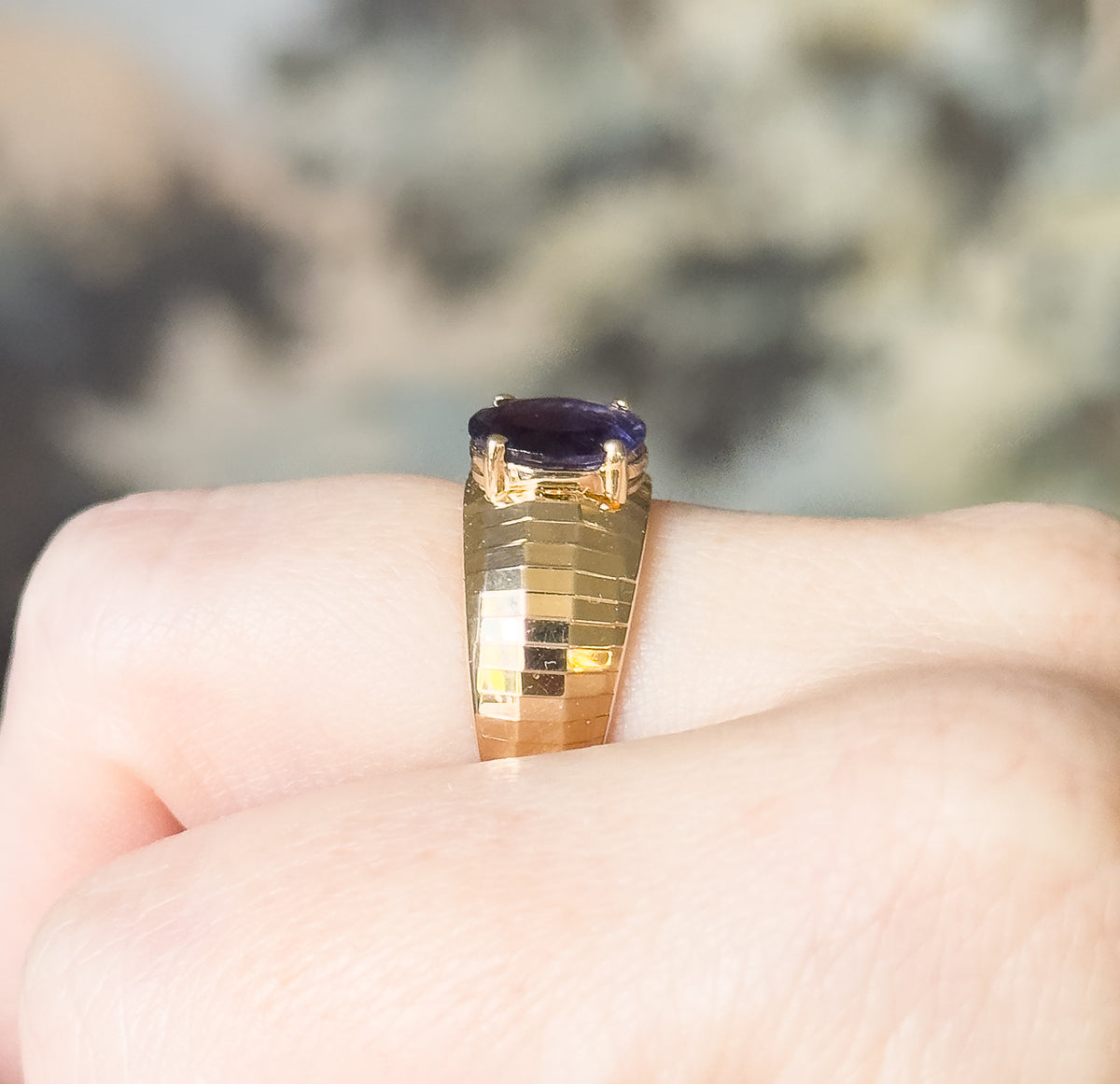 Head-on view along the finger, showing the four prongs gripping the oval iolite and the stepped facets of the yellow gold band catching light as they taper down each side.