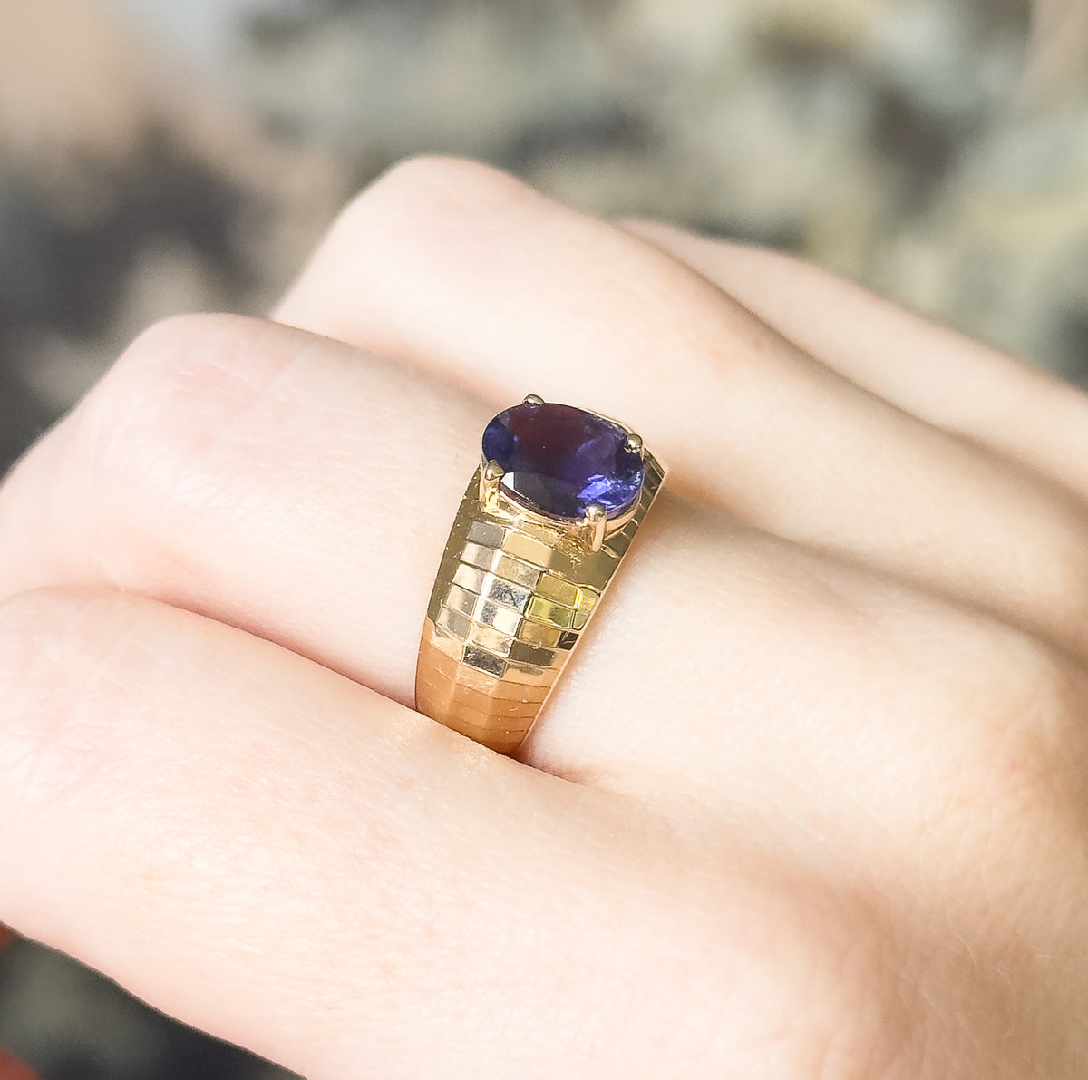 Angled close view from the side, the deep bluish purple oval iolite sitting high on its prongs above the checkered facets of the broad yellow gold band, shown on a finger.