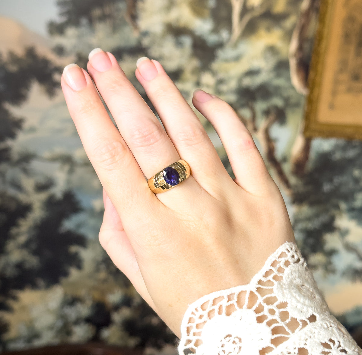 The back of the hand raised against a leafy painted backdrop, the ring on the middle finger with the iolite reading as a small dark violet oval on its faceted yellow gold band.
