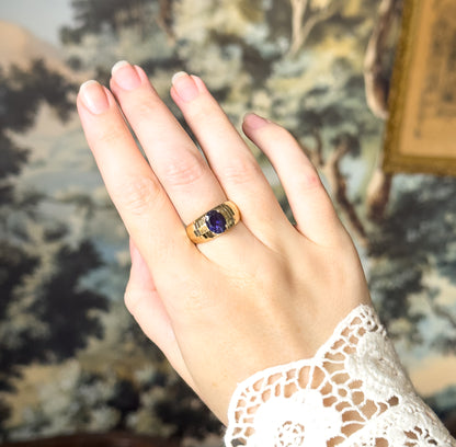 The back of the hand raised against a leafy painted backdrop, the ring on the middle finger with the iolite reading as a small dark violet oval on its faceted yellow gold band.