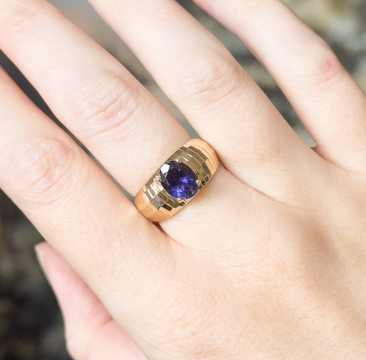 An iolite solitaire ring in 10K yellow gold, shown on a finger, the oval bluish purple stone prong-set in a wide faceted band that narrows toward the shank.