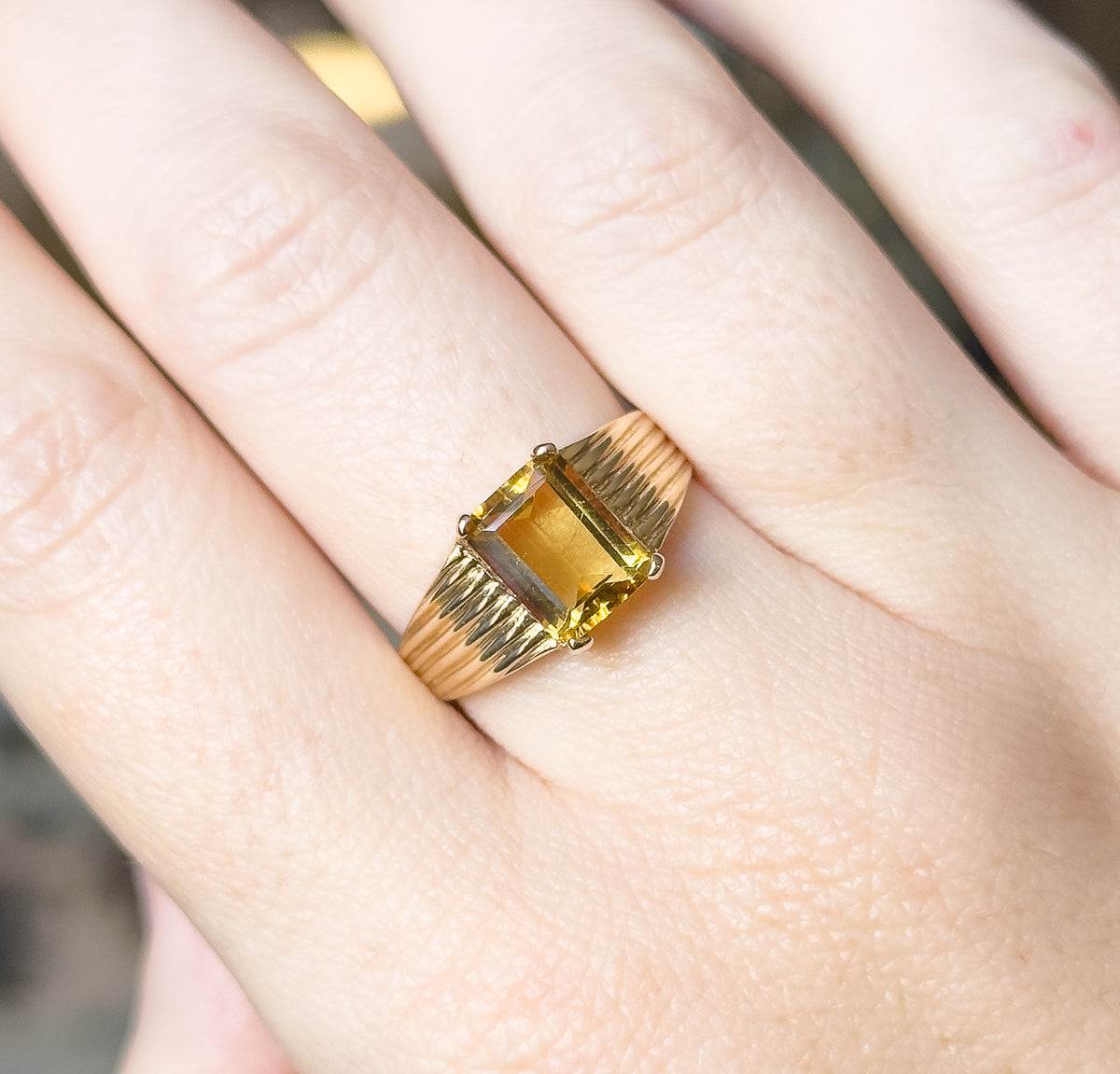 Shown on a finger from above and close in, the golden emerald-cut stone held at its corners by four prongs, with the ribbed shoulders flaring either side of the raised setting.