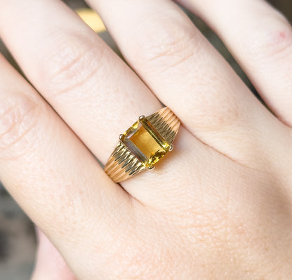 Shown on a finger from above and close in, the golden emerald-cut stone held at its corners by four prongs, with the ribbed shoulders flaring either side of the raised setting.