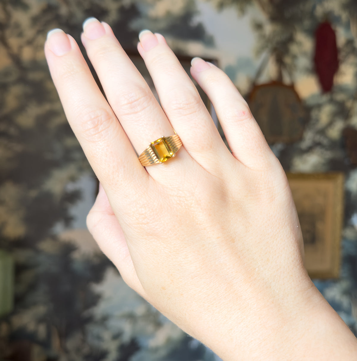 The whole hand is shown at a distance against a soft painted backdrop, the ring small on the ring finger, giving a sense of its modest scale.