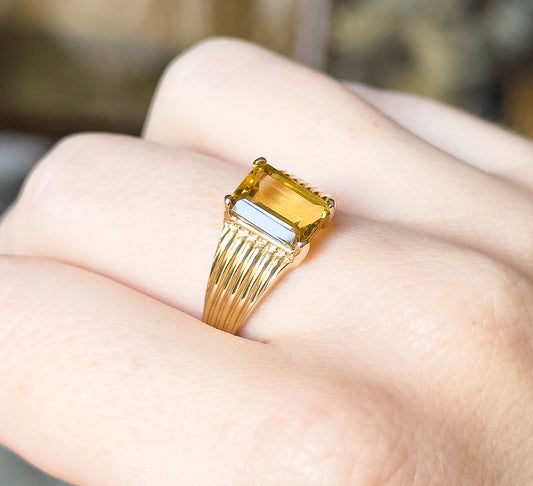 An emerald-cut citrine ring in 10 karat yellow gold, shown on a finger, the prong-set golden stone raised above ribbed shoulders that taper to a slim shank.