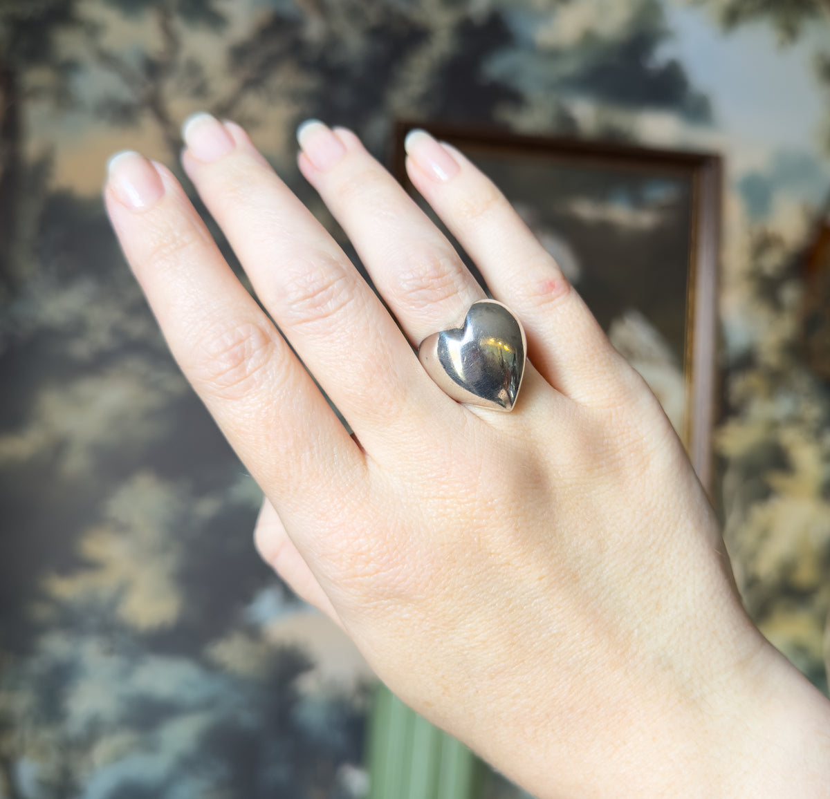 The whole hand is shown against a leafy patterned backdrop, the puffy heart sitting on the ring finger where its domed face spans much of the finger's width.