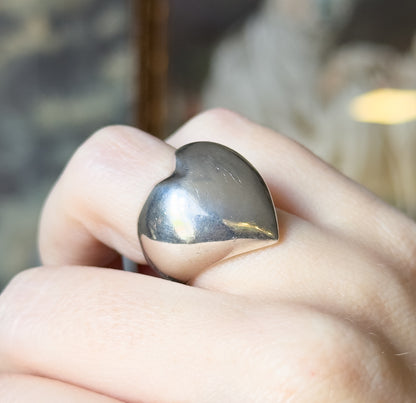 Sterling silver puffy heart ring, shown on a finger in close-up, the rounded heart face angled across the knuckle above a narrow tapering shank.