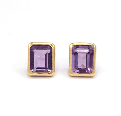 Almost the same view with slightly different angle, cropped closer so the cut corners of each yellow gold bezel and the broad central facet down each purple amethyst fill the frame.