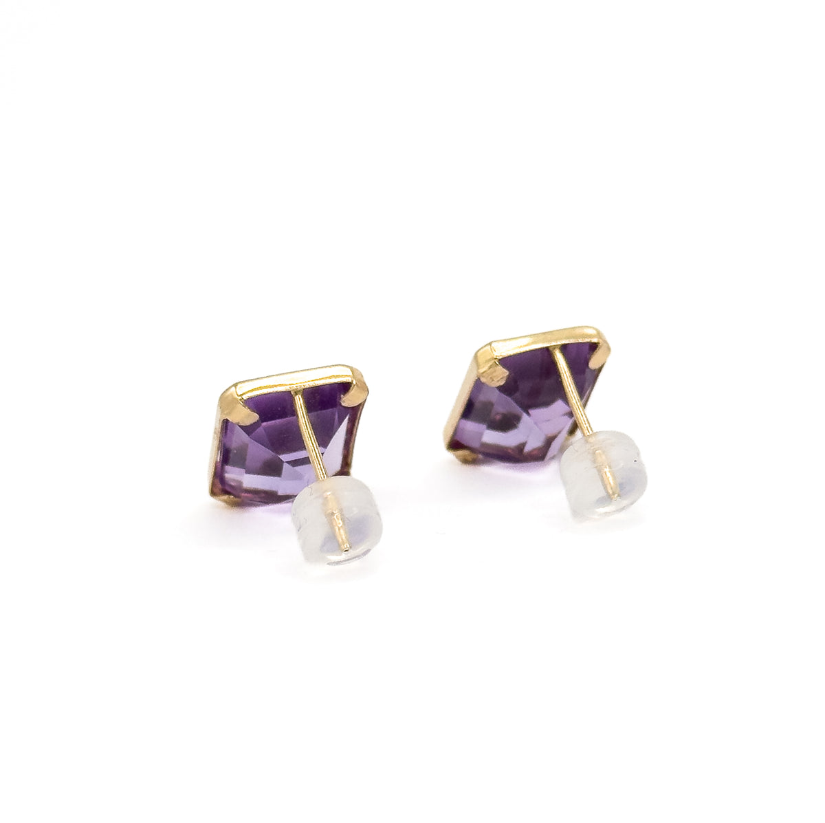 Both studs angled towards the viewer against white, the yellow gold bezel corners catching the light above the purple stones, with each post and its clear backing pushed close beneath.