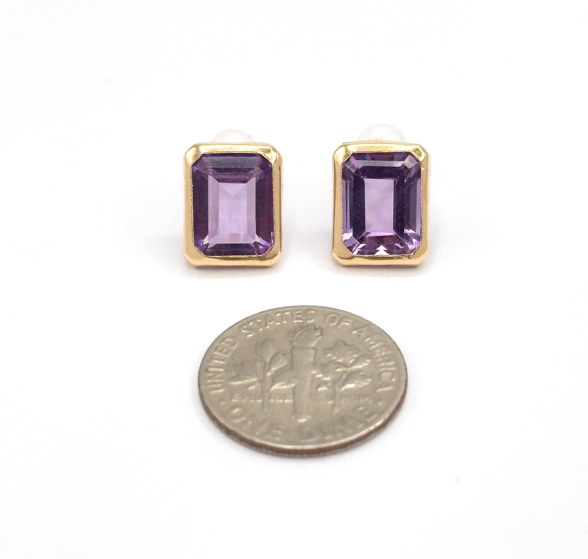 Both studs face-up above a dime, the coin showing how the yellow gold bezels and purple emerald-cut amethysts measure barely more than half its width.