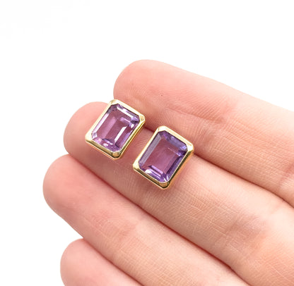 Emerald-cut amethyst stud earrings in 14K yellow gold bezels, shown resting on a fingertip against white, the soft purple stones each about the width of a fingernail's edge.