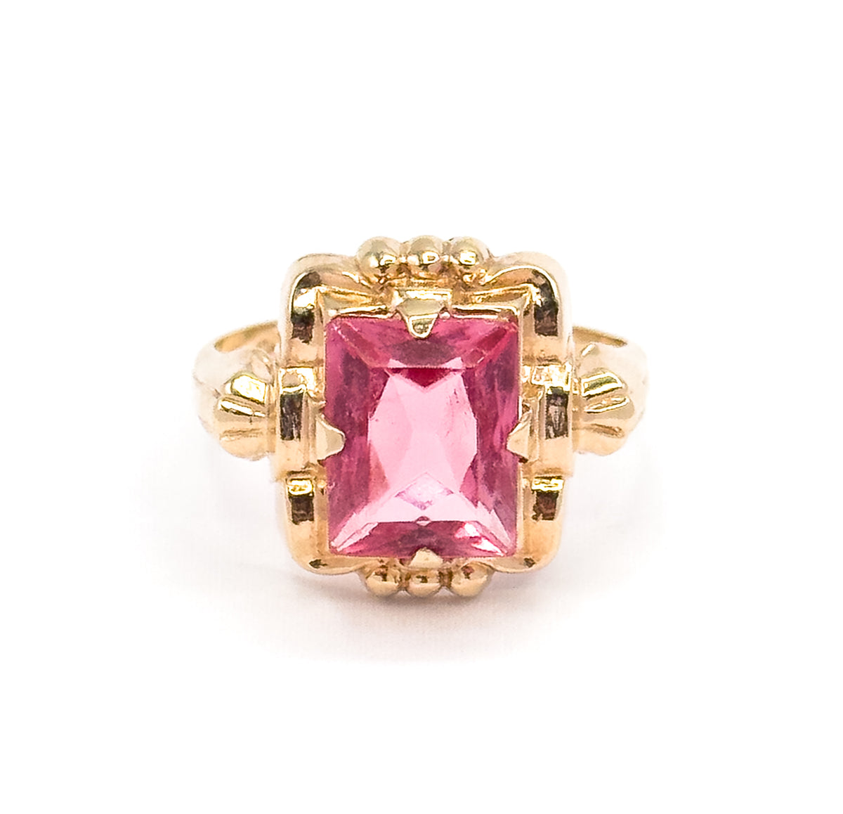 Straight-on from the front, the rectangular radiant-cut pink glass fills the squared yellow gold frame, flanked by beaded accents above and below and flared shoulders at each side.