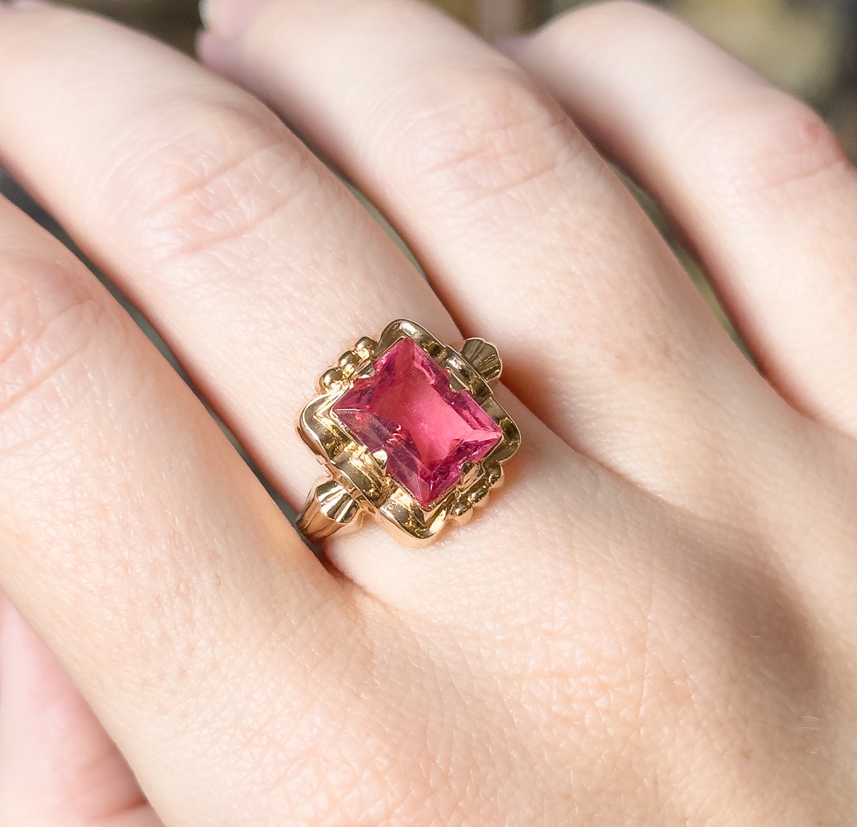 Shown on the hand at a closer angle, the pink glass stone catches light across its facets while the scalloped yellow gold flanks and shoulders sit low against the finger.