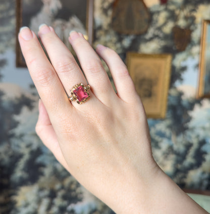 The whole back of the hand fills the frame against a wall of framed pictures, the pink glass ring on the ring finger showing how small the face sits against the knuckles.