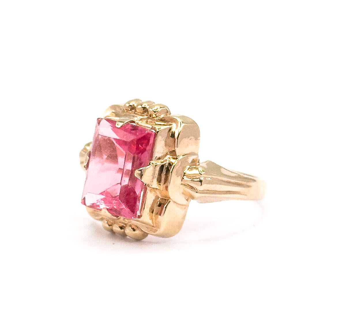 Angled three-quarter view with the shank receding to the right, catching the stepped facets across the face of the rectangular pink glass and the beaded yellow gold frame around it.