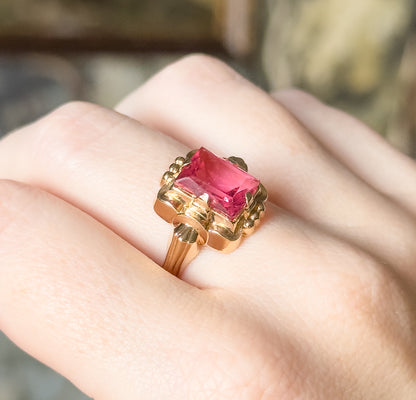 Mid-century ring with a rectangular radiant-cut pink glass stone in 10K yellow gold, shown on a finger, its scalloped bezel spreading wide before tapering into a slim shank.