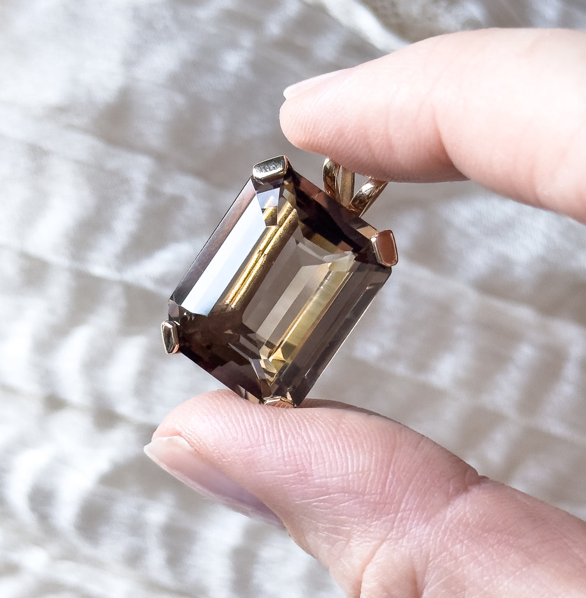 A smoky quartz pendant in 10K yellow gold, the emerald-cut golden brown stone held by four prongs with its bail above, held between two fingertips that show its size.