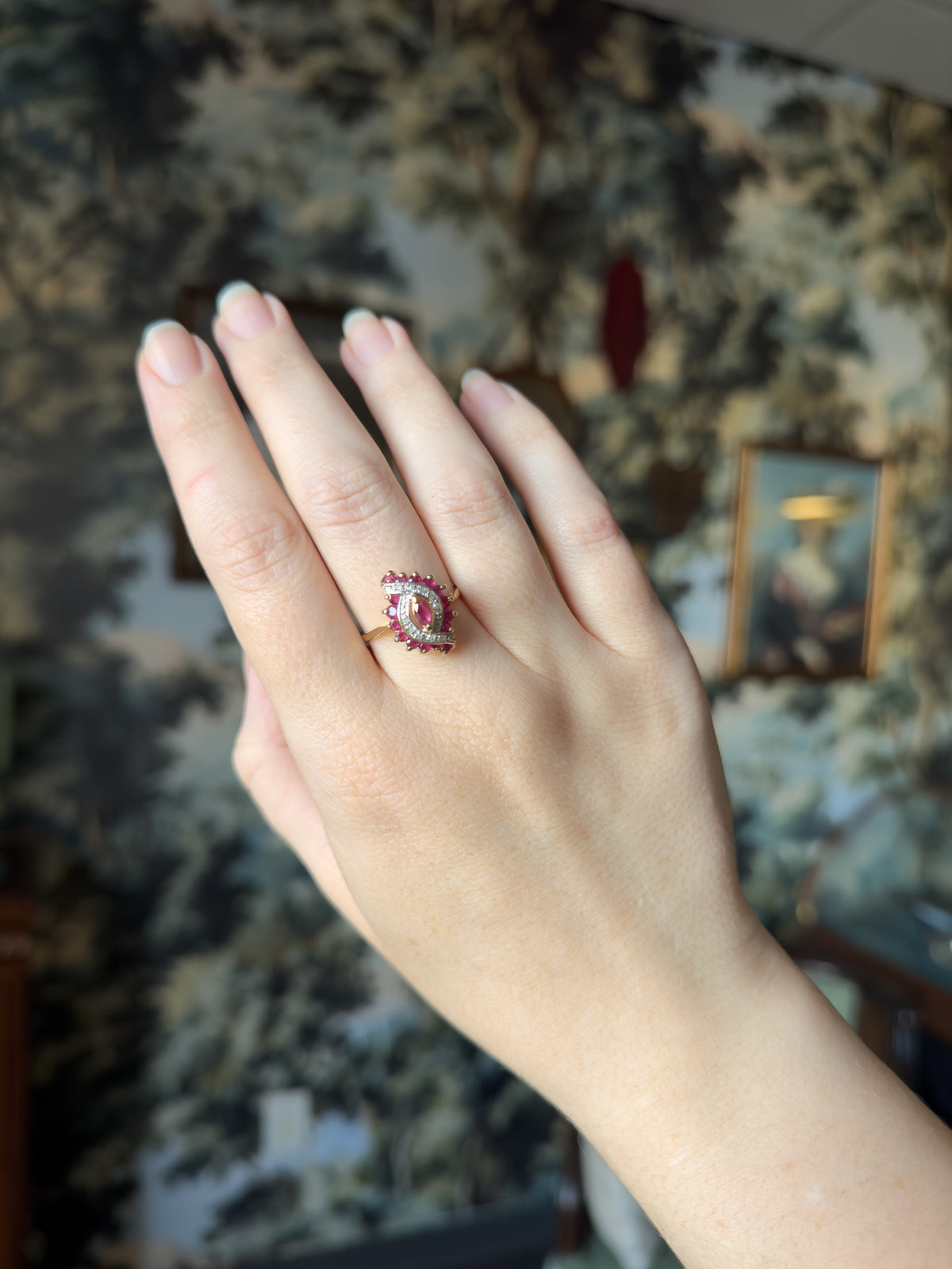 The whole hand held upright against a leafy patterned wall, the ring small on the finger so its marquise face reads as little wider than the finger itself.