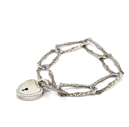 Laid flat on a white background and loosely coiled, the open elongated links show their milgrain edges and floral engraving, with the heart-shaped lock clasp and its keyhole facing forward.