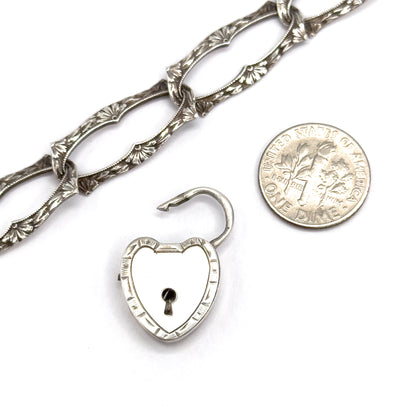 The heart lock with its shackle open lies beside a dime for scale, the lock about the same size as the coin, with the bracelet's floral links above.