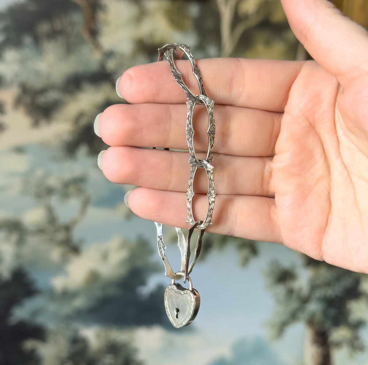 An Edwardian sterling silver bracelet of elongated floral links, draped over open fingers with its heart-shaped lock clasp hanging below, the hand giving a sense of scale.