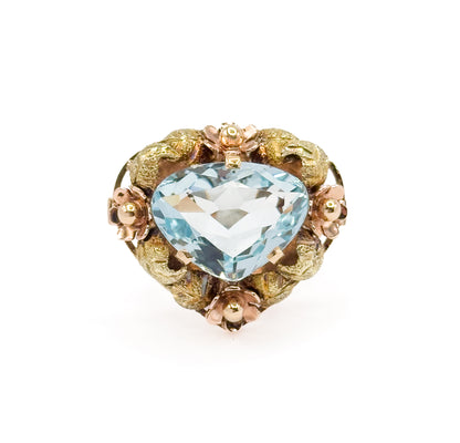 Straight-on view of the face alone, the icy blue aquamarine prong-set at the centre with rose gold flowers at top, bottom and both sides between green gold leaves, the shank hidden behind.