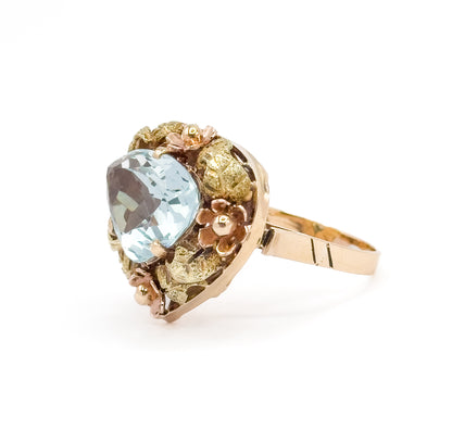 Near-profile from the stone's point, the icy blue aquamarine seen edge-on to show its depth while rose gold flowers step around the rim and the polished shank runs away to the right.