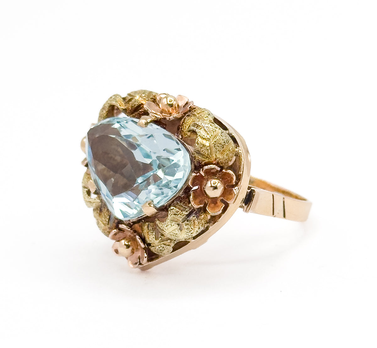 Raised three-quarter view from the stone's side, the open pierced gallery visible beneath the green gold leaves and rose gold flowers as the shank angles back to the right.