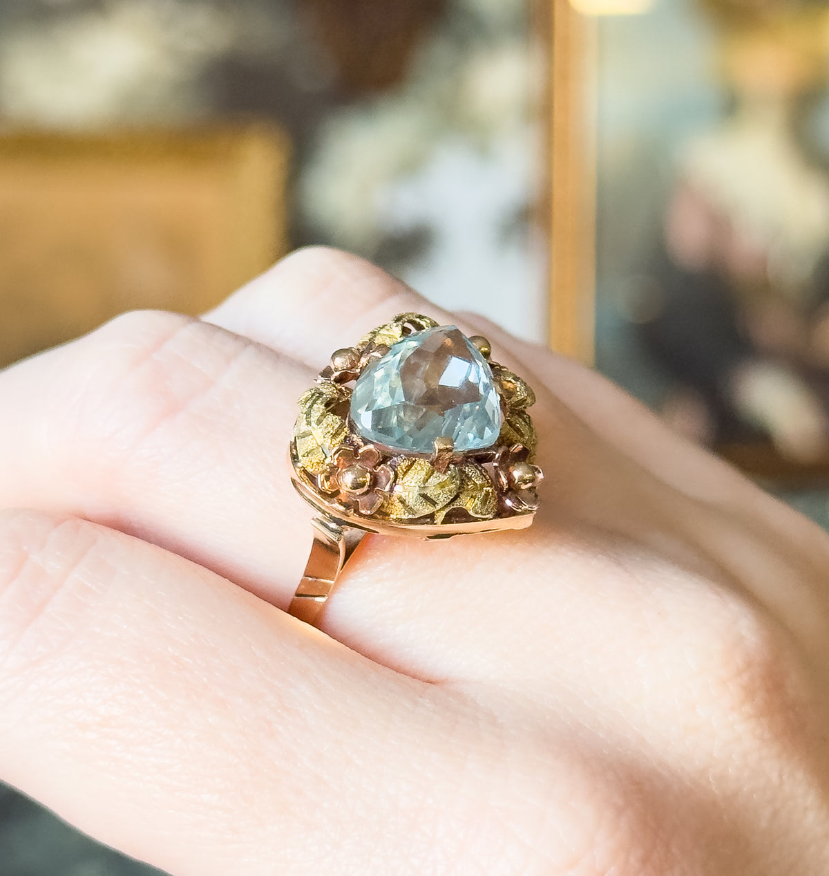 Vintage aquamarine floral cocktail ring in 18K two-tone gold, shown on a finger, its icy blue prong-set stone framed by green gold leaves and rose gold flowers above a tapering shank.