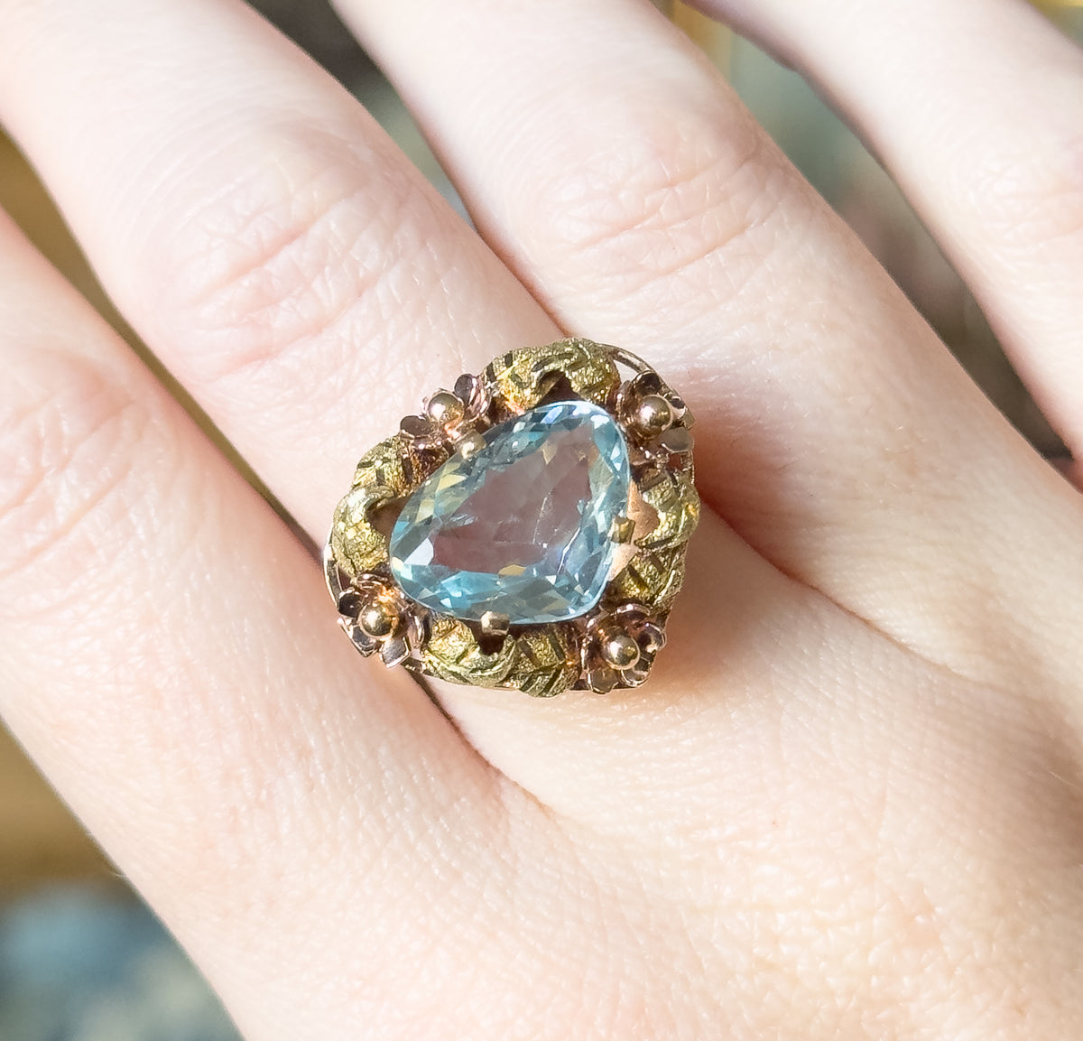Overhead view down onto the ring on the finger, showing the full triangular outline of the icy blue stone and the ring of leaves and flower heads encircling it.