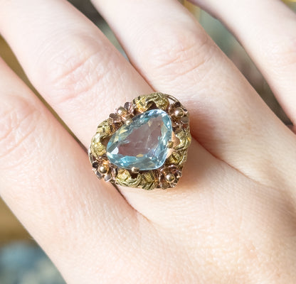 Overhead view down onto the ring on the finger, showing the full triangular outline of the icy blue stone and the ring of leaves and flower heads encircling it.