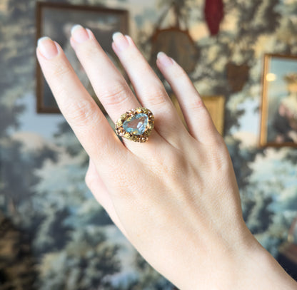 The whole back of the hand shown against a painted scenic backdrop, giving a sense of the ring's scale as it sits across the finger with its floral face raised.