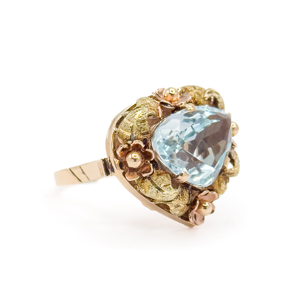 Angled three-quarter view against white, the icy blue aquamarine catching light across its facets while rose gold flowers and green gold leaves ring the bezel and the shank sweeps away to the left.