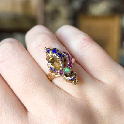 Shown on a finger from above with the hand relaxed, the face tilted across the knuckle so the opal flashes green beside the row of small rubies curving up the blue enamel edge.