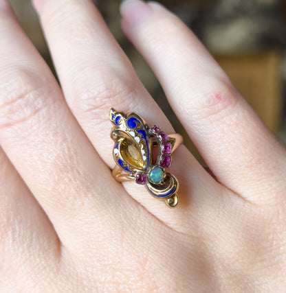 Shown on a finger with the hand turned so the face angles away, the opal flashing green at the lower tip, the five rubies in a line beside the blue and white enamel scrolls.
