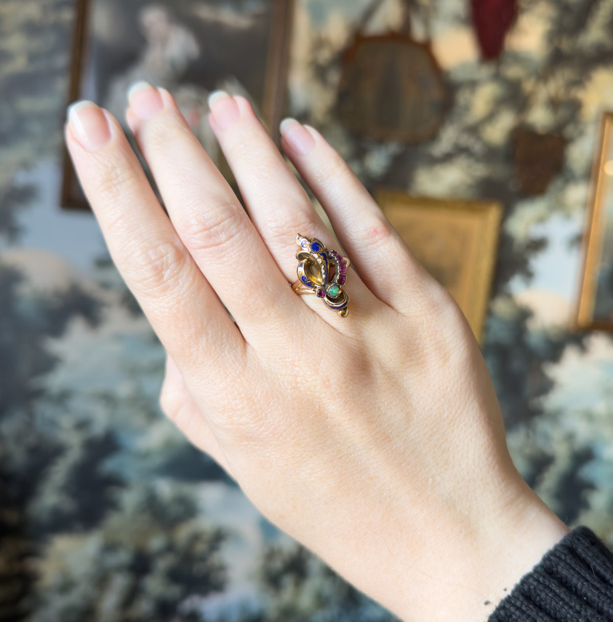 Shown on a finger with the whole hand laid flat and fingers together, the small face reading as a bright cluster of blue enamel, rubies and green-flashing opal against the broad back of the hand.