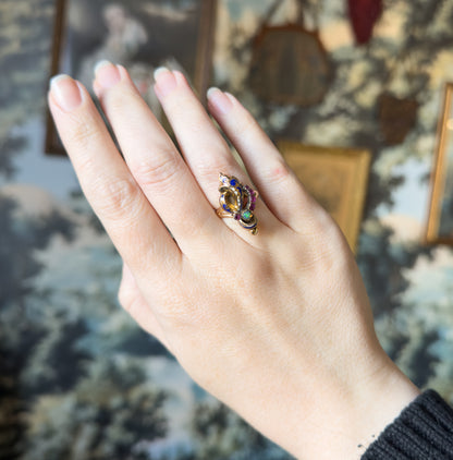 Shown on a finger with the whole hand laid flat and fingers together, the small face reading as a bright cluster of blue enamel, rubies and green-flashing opal against the broad back of the hand.