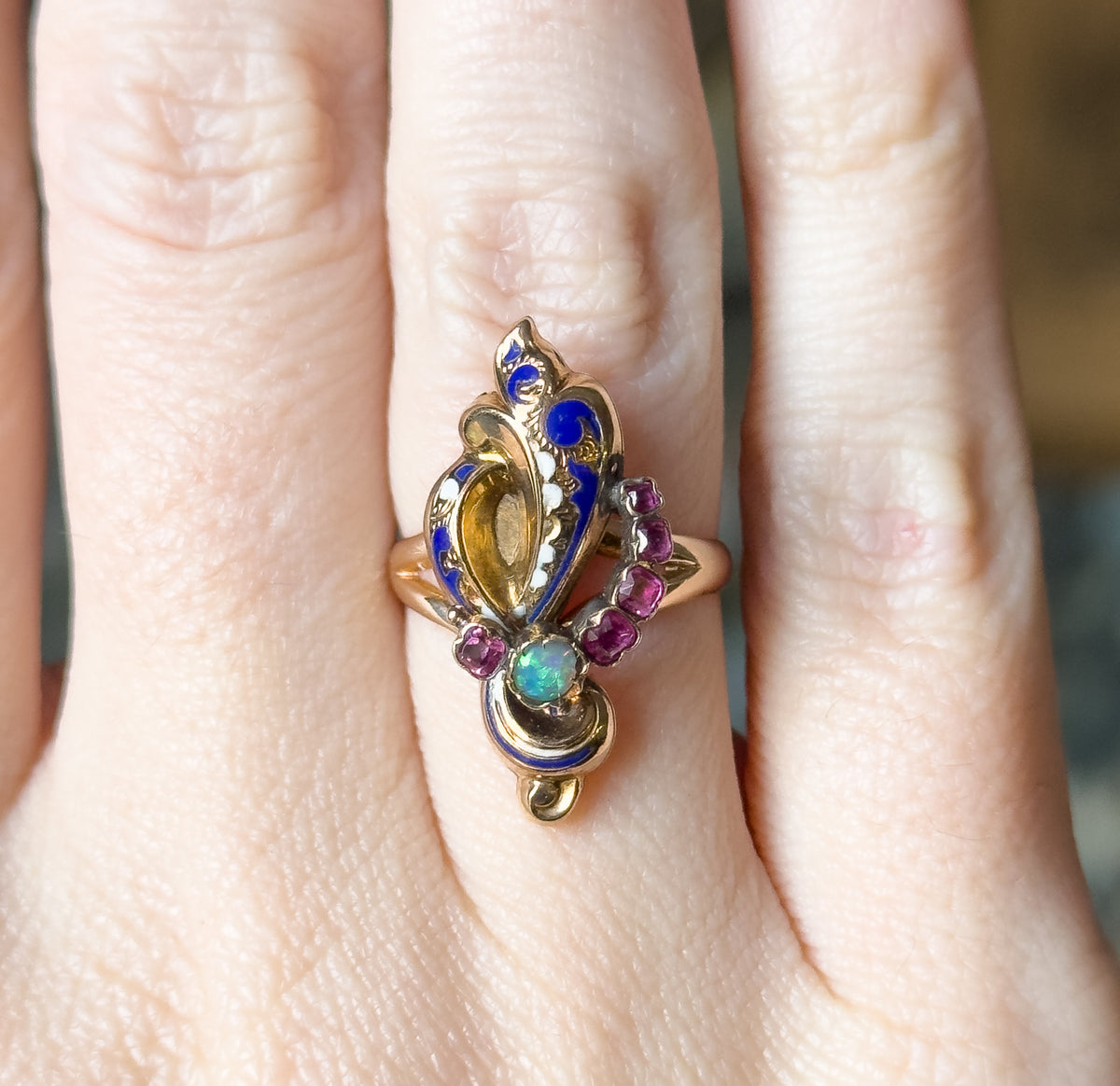 Shown on a finger, the ring's long face lies lengthwise along the knuckle, giving its scale against the hand, with the opal and rubies catching light at the lower end.